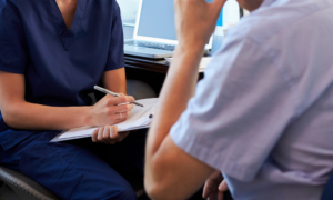 A nurse writes notes on a clipboard while person with their back to camera talks.