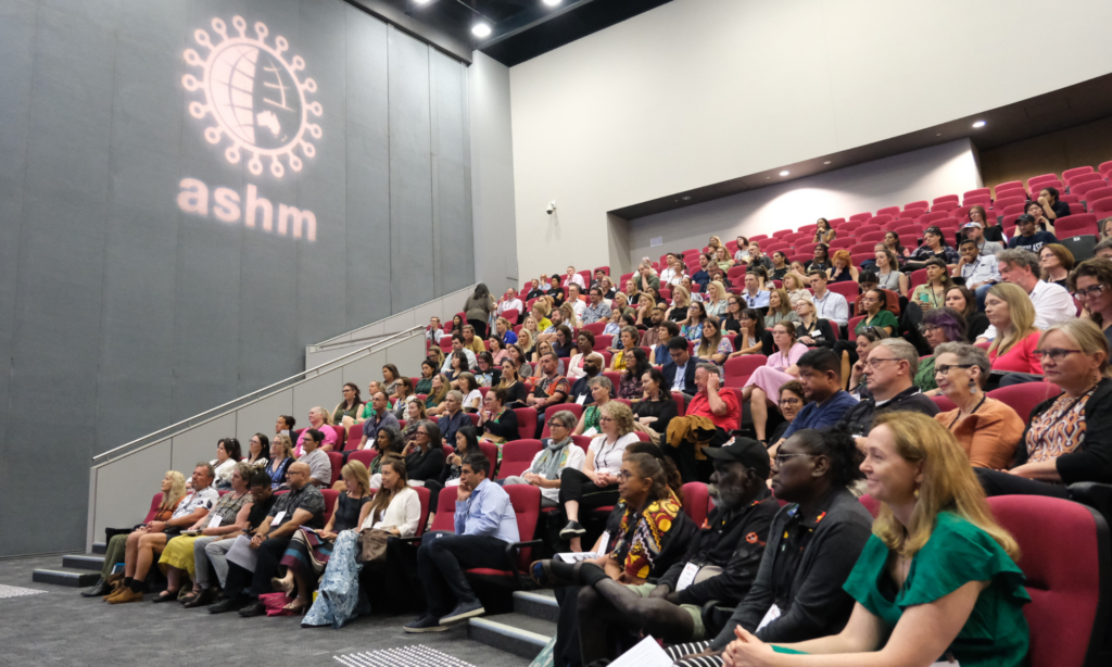 Crowd of people sitting an auditorium with ASHM logo light above.
